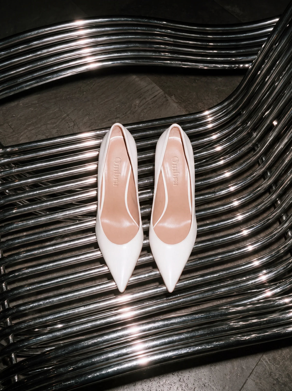 White pointed heels with minimal branding using Silk Sans typography, photographed on sculptural chrome furniture.