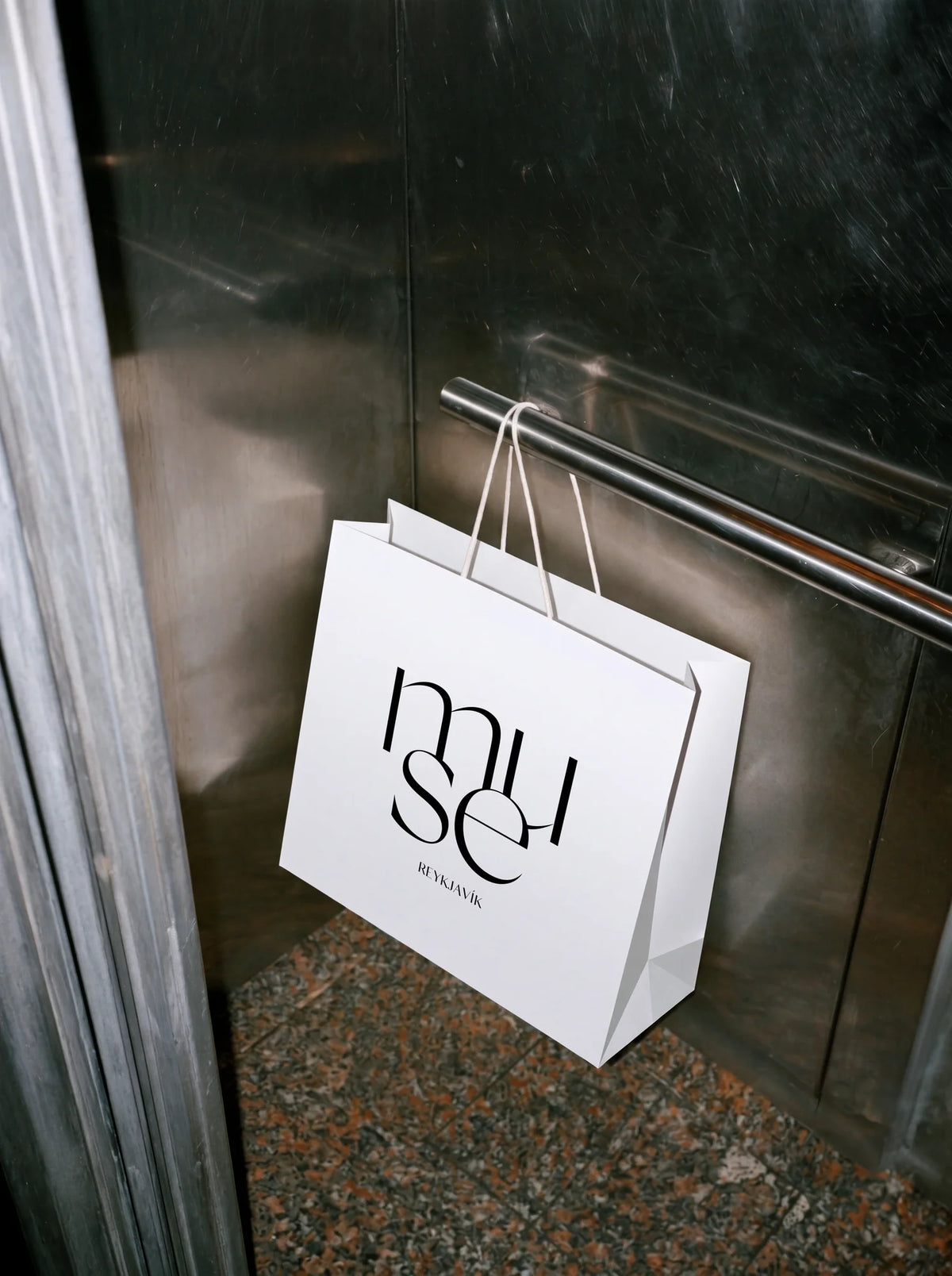 White shopping bag with Muse Reykjavík branding and custom Silk Sans typography hanging inside a metal elevator.
